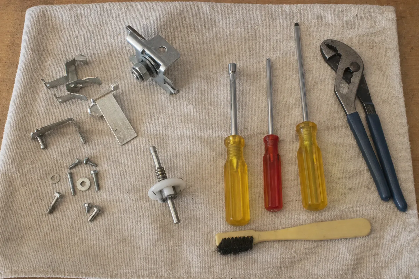 Stainless clips and a flat blade bracket beside a chrome pivot assembly and a shaft with a white collar and spring, with nut drivers, two screwdrivers, pliers and a small brush on a cream cloth