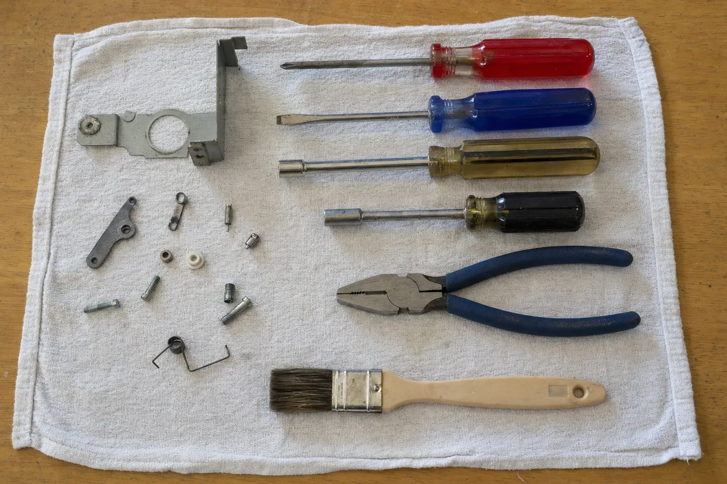 Stamped steel bracket with a large round cut-out, a small cast lever, a wire spring, nylon and metal bushings and screws on a white towel, with four drivers, pliers and a soft brush