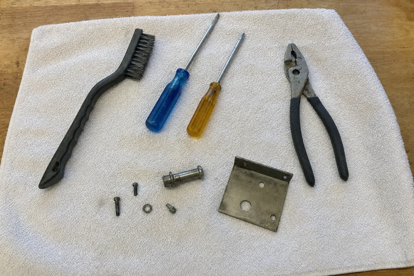 Black-handled wire brush, a cross-head and a flat screwdriver, slip-joint pliers, a shouldered pin, a nut, three screws and a drilled steel plate on a white towel